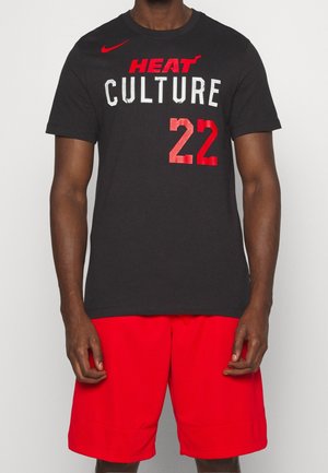 Mężczyzna noszący czarną koszulkę z czerwonym napisem "HEAT CULTURE 22" i czerwonym logo Nike, w połączeniu z jasnoczerwonymi szortami.