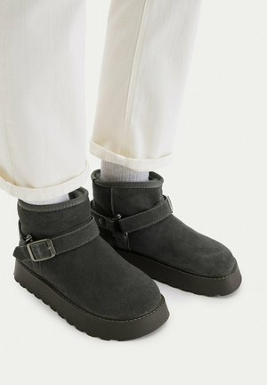 Black suede ankle boots with buckle straps worn with white socks and rolled-up white pants on a white background.