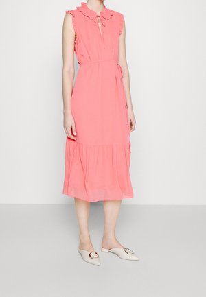 Kobieta w koralowej sukience midi bez rękawów z falbankowym kołnierzem oraz beżowymi wsuwanymi butami na płaskim obcasie z ozdobnymi klamrami.