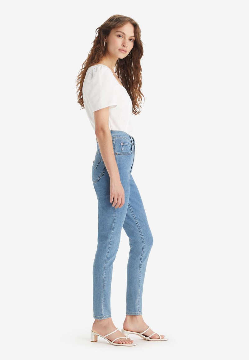 Jean taille haute bleu clair avec une coupe slim, fabriqué en denim. Associé à un haut blanc à manches courtes et à des sandales à talons blanches.