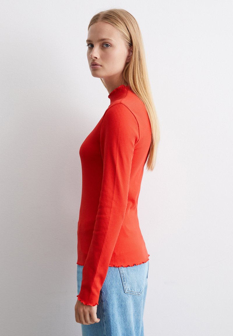 Rotes geripptes Langarmoberteil mit hohem Halsausschnitt und fransigen Abschlüssen, kombiniert mit hellblauen Jeans. Der Stoff hat eine strukturierte Optik.