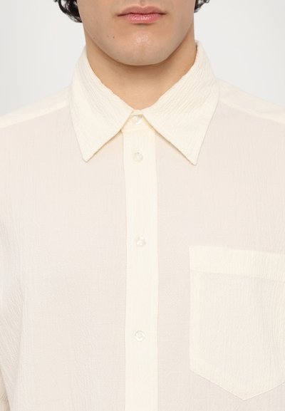 Chemise boutonnière de couleur crème avec un tissu texturé, dotée d'un col classique, de cinq boutons et d'une poche avant sur le côté gauche.