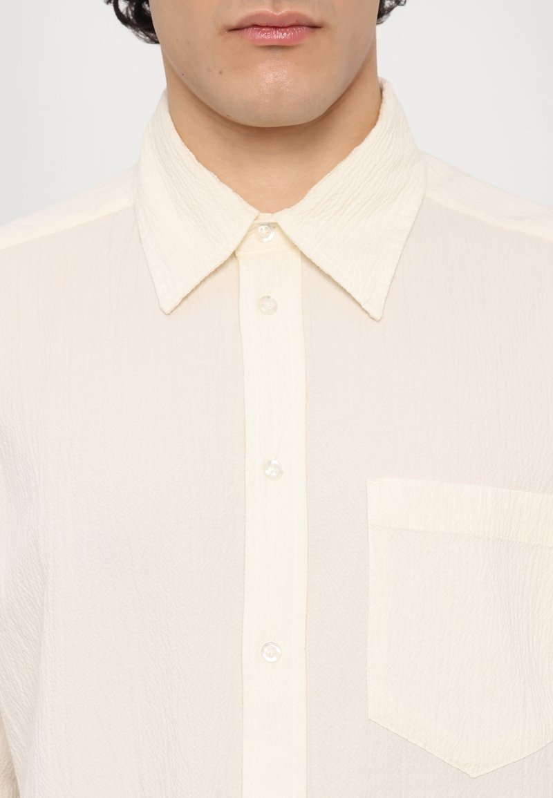 Camisa abotonada color crema con una tela texturizada, que presenta un cuello clásico, cinco botones y un bolsillo frontal en el lado izquierdo.