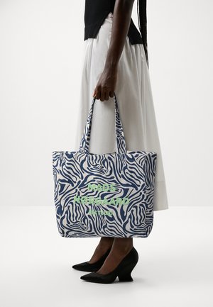 Sac fourre-tout en tissu recyclé avec motif zèbre bleu marine et blanc, comportant le texte vert "MADS NØRGAARD" sur le devant. Les anses sont solides et assorties.