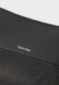 Čierny textúrovaný clutch s geometrickým vzorom, hladkou hornou časťou a embosovaným bielym logom "Calvin Klein".