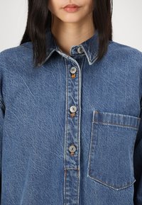 Denim srajca v srednje modri barvi z gumbom spredaj, koničastim ovratnikom in enim žepom na prsih. Ima srebrne gumbe in vidno šivanje.