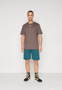T-shirt de algodão castanho com um logotipo amarelo, combinado com calções turquesa. O visual é complementado com ténis de cano alto em preto e bege.