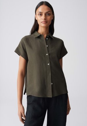 Frau trägt kurzärmliges dunkelolivfarbenes Hemd mit Knöpfen und schwarze Hose, steht vor einem schlichten hellgrauen Hintergrund.