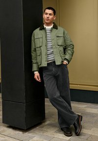 Grüne Nylon-Bomberjacke mit utility Taschen, kombiniert mit gestreiftem Strickpullover und weit geschnittenen, dunkelgrauen Hosen, ergänzt durch schwarze Schuhe.