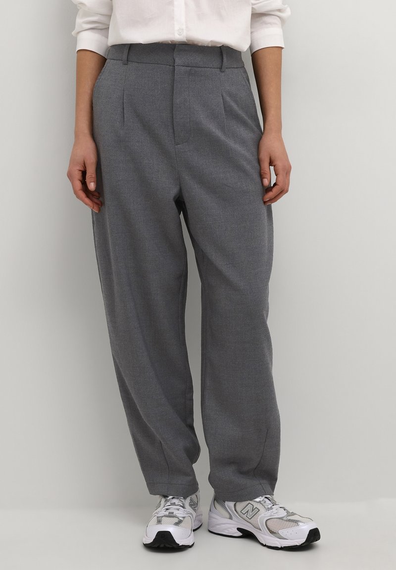 Personne portant un pantalon gris ample, des baskets blanches et une chemise blanche à manches longues, debout devant un mur clair uni.