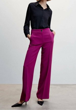 Vrouw draagt een zwarte blouse met lange mouwen, hooggetailleerde wijde magenta broek, en zwarte pumps met puntige neus, staand met handen in de zakken.