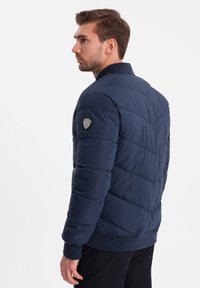 Námořnická modrá péřová bunda s šikmým vzorem, žebrovaným límcem a logem na levém rameni. Texturovaný povrch, zapínání na zip.