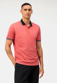 Polo rose avec un col noir présentant des rayures blanches et bleues, manches courtes, et un petit logo sur la poitrine. Pantalon noir.
