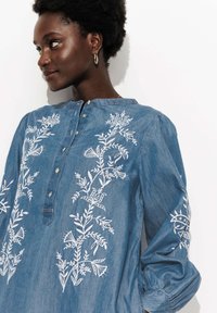 Femme aux cheveux courts et bouclés portant une blouse bleue à boutons avec une broderie florale blanche, regardant à gauche sur un fond blanc.