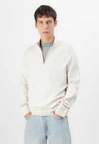 Beige Viertelreißverschluss-Pullover aus weichem Stoff, mit gerippten Bündchen und Saum, über einem hellgrauen Hemd getragen, kombiniert mit hellen Jeans.