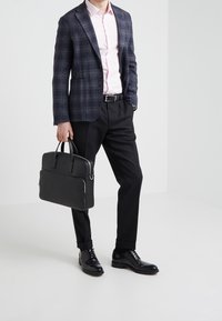Maletín negro con dos asas, llevado con un blazer azul marino de cuadros y pantalones negros. Camisa rosa claro, zapatos negros pulidos y cinturón negro.