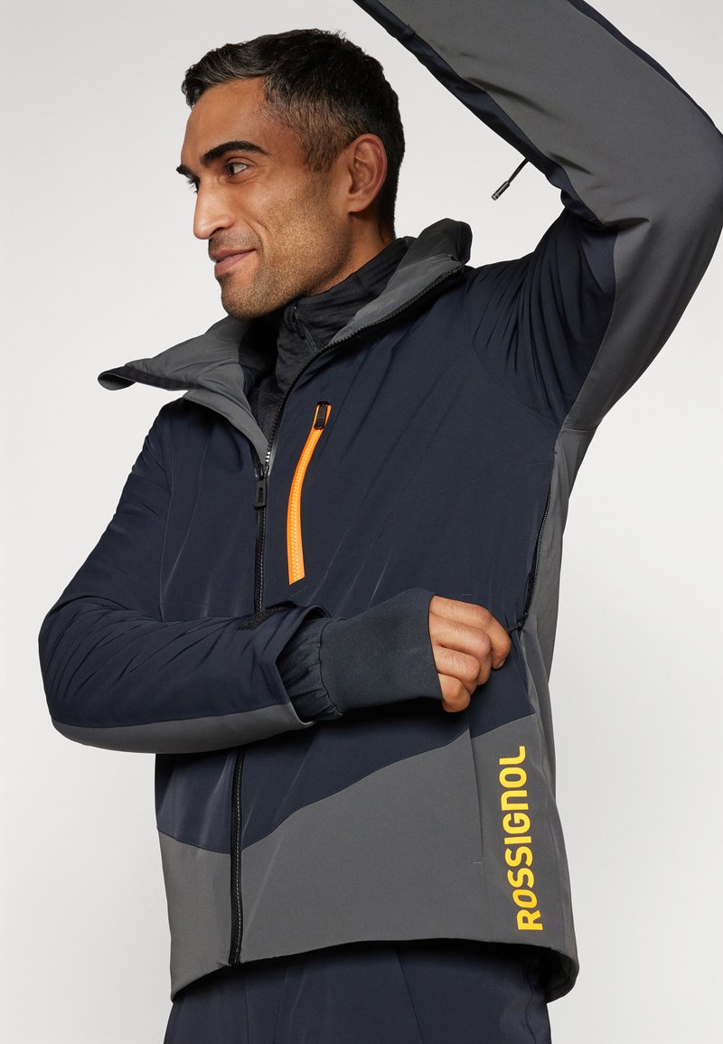 Giacca con cappuccio blu navy e grigia con una tasca con zip arancione, aperture per i pollici sui polsini e il logo "ROSSIGNOL" in giallo sull'orlo.