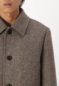 Cappotto marrone con texture, colletto a punta, caratterizzato da bottoni neri e un sottile motivo a chevron. Vista ravvicinata della parte superiore anteriore.