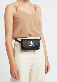 Černá kožená pásková taška s texturovaným povrchem, stříbrnou logo sponou a kompaktním obdélníkovým tvarem, nošená přes světle barevné kalhoty.