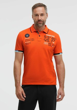 Mann mit kurzen Haaren und Bart, der ein orangefarbenes Poloshirt mit schwarzem Text und Emblemen trägt, steht vor einem weißen, schlichten Hintergrund.