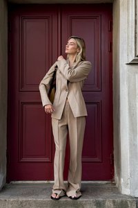 Beżowy garnitur o dopasowanym kroju z długimi rękawami i jednym guzikiem, w zestawieniu z luźnymi spodniami. Stoi na kamiennym stopniu przed czerwonymi drzwiami. Na nogach sandały.