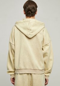 Felpa beige con una vestibilità rilassata, maniche oversize, cappuccio con cordino e orlo a coste. Materiale in pile morbido con una texture liscia.