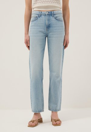 Mujer con jeans rectos de talle alto en azul claro y sandalias de tacón beige, de pie contra un fondo liso y claro.