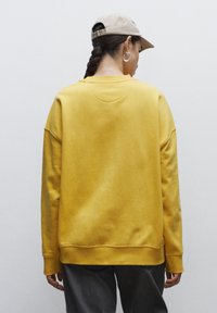 Sudadera amarilla con un ajuste relajado, puños y dobladillo acanalados, textura suave. El modelo lleva una gorra beis y pantalones grises. Vista desde la parte posterior.