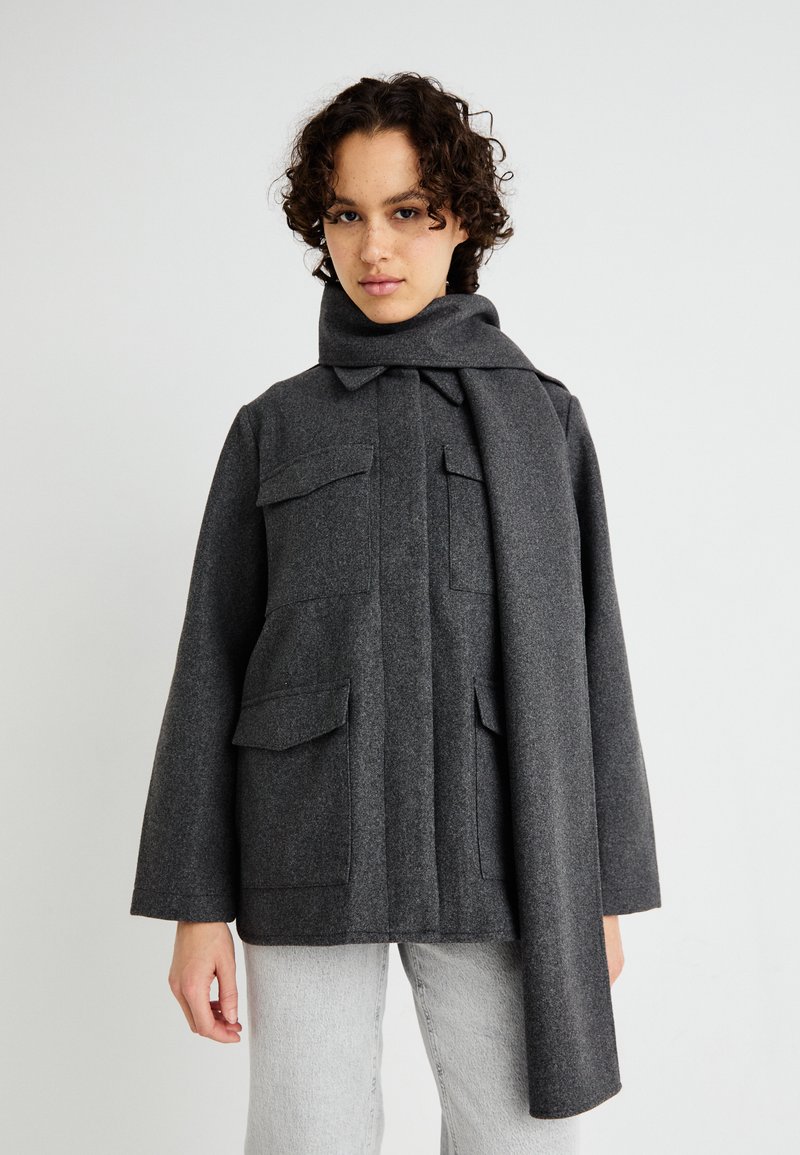 Cappotto grigio doppiopetto con colletto alto, pattina e quattro tasche frontali. Realizzato in tessuto testurizzato con una sciarpa attaccata.