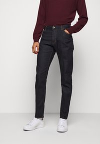 Person som bär mörka slim-fit jeans, burgundy långärmad topp och vita sneakers, står med en hand i fickan mot en enfärgad bakgrund.