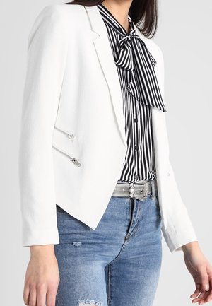 Frau trägt einen weißen Blazer mit silbernen Reißverschluss-Details über einer schwarz-weiß gestreiften Bluse mit Schleife, kombiniert mit ausgewaschenen blauen Jeans und einem silbernen Gürtel.