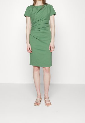 Kobieta w zielonej sukience za kolano z krótkimi rękawami i marszczeniem po bokach, ubrana w białe sandały odkrywające palce, stojąca na jednolitym białym tle.