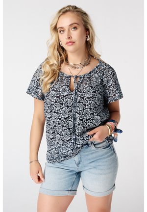 Vrouw met lang blond haar draagt een zwart-wit bloemenblouse, lichte spijkershort, gelaagde kettingen en armbanden met een gebonden stoffen band.