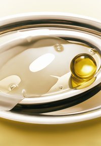Okrągła, odbijająca światło taca z metaliczną krawędzią; przezroczysta żelowa kapsułka w żółtym kolorze oraz krople oleju spoczywające na gładkiej, jasnej powierzchni.