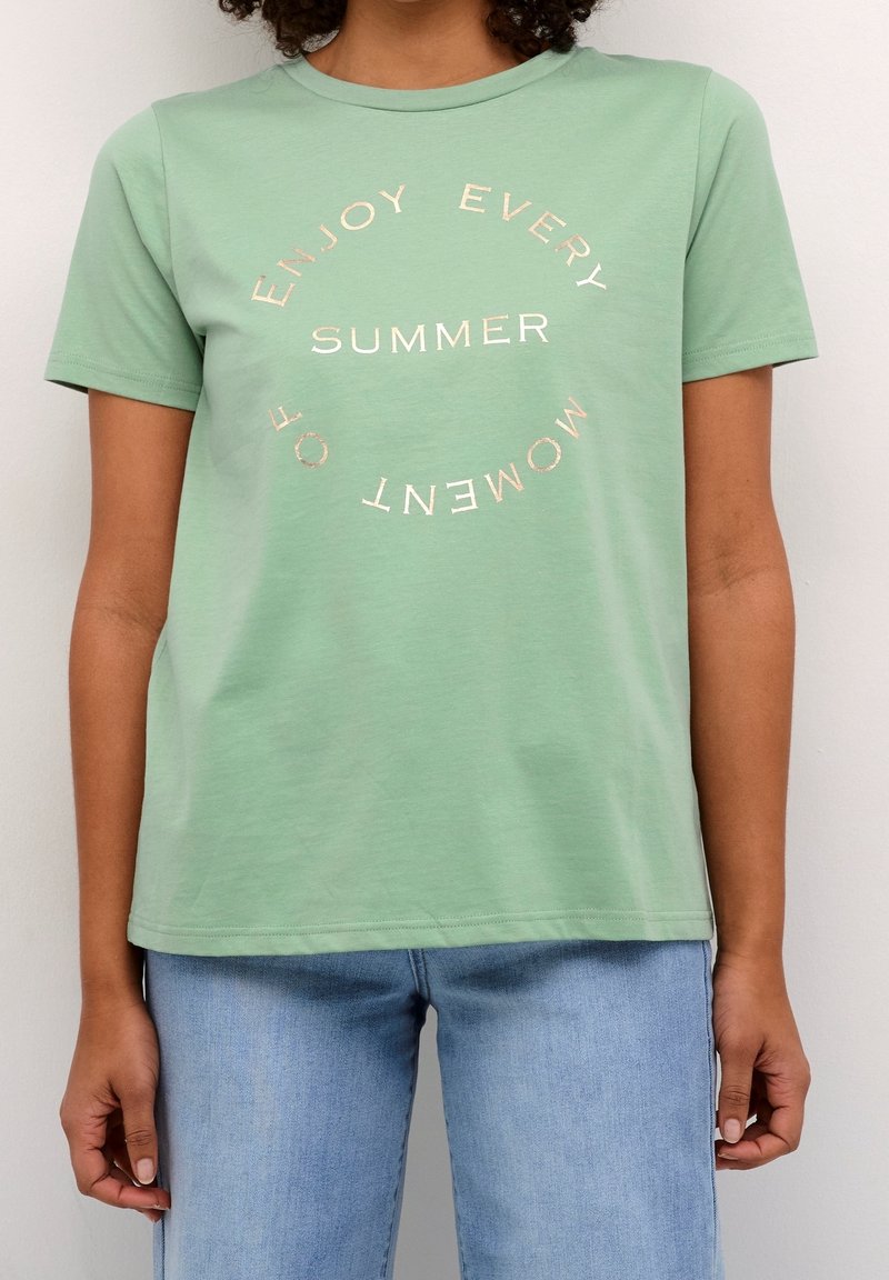 T-shirt en coton vert clair avec des manches courtes et un col rond. Présente un texte doré qui dit "SAVOUREZ CHAQUE MOMENT DE L'ÉTÉ."