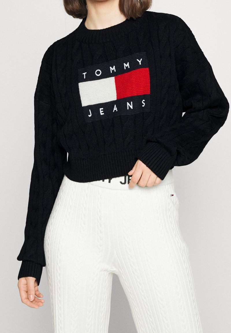 Femme portant un pull court noir en maille torsadée avec le logo « Tommy Jeans » et un pantalon blanc en maille torsadée taille haute.