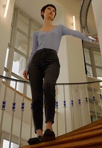 Top de manga longa azul claro com nervuras, jeans pretos de cintura alta e sapatilhas pretas. O sujeito está em uma escada de madeira com corrimão.