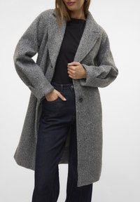 Manteau en laine gris avec un col en V, des manches longues et une coupe décontractée. Il présente une fermeture à un bouton et une surface texturée. Porté par-dessus un jean sombre.