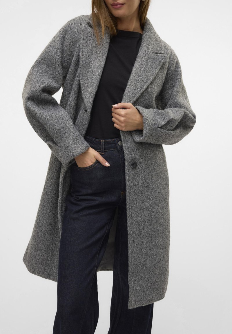 Manteau en laine gris avec un col en V, des manches longues et une coupe décontractée. Il présente une fermeture à un bouton et une surface texturée. Porté par-dessus un jean sombre.