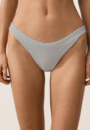 Femme portant un bas de bikini blanc avec un petit motif ovale noir, vue de la taille jusqu'au milieu des cuisses sur un fond uni.