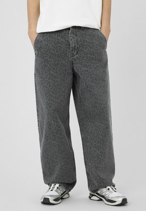 Personne portant un jean à jambes larges, noir délavé à imprimé léopard, les mains dans les poches, ainsi que des baskets argentées sur un fond gris uni.