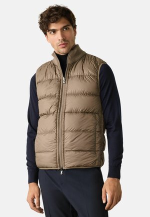 Mann trägt beige gesteppte ärmellose Weste über schwarzem Langarmshirt und steht vor schlichter, heller Hintergrund.