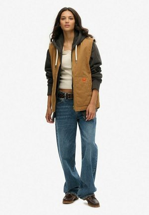 Chaleco acolchado marrón con una sudadera con capucha gris oscuro por debajo, jeans de pierna ancha azules y zapatos marrones. El chaleco tiene un bolsillo delantero y cierre de cremallera.