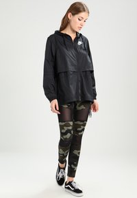 Svart lättviktig vindjacka med huva, med dragkedja och Nike-logotyp; kombinerad med camo nätleggings och svarta sneakers.