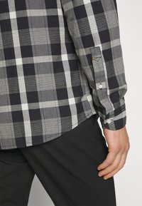 Černobílá kostkovaná košile s manžetou na knoflíky, s různými vzory kostkované látky a hladkou texturou. Kombinována s černými kalhotami.