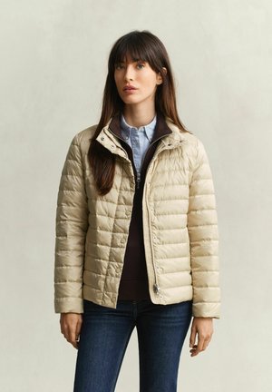 Woman with long brown hair wearing a beige quilted jacket, dark zip-up sweater, light blue shirt, and dark blue jeans against a plain background.