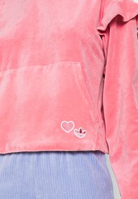 Sudadera corta de terciopelo rosa con detalle de mangas con volantes, que presenta un bolsillo frontal y acentos bordados de corazón y logotipo en el dobladillo.