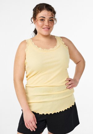 Kvinde med krøllet hår og fregner iført en gul ærmeløs top med blonder og sorte shorts, stående med den ene hånd på hoften.