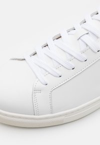 Weißes Leder-Sneaker mit flachen weißen Schnürsenkeln, glatter Textur, durchbrochener Seitendetailierung und einer Gummisohle mit strukturiertem Griffmuster.
