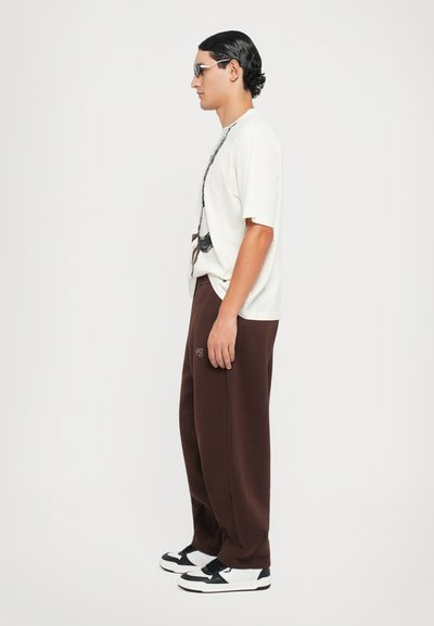 White graphic t-shirt, relaxed fit, paired with brown sweatpants featuring side pockets and purple branding. Completed with black and white sneakers.